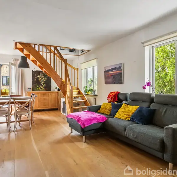 Grå sofa med farverige puder står foran store vinduer med grøn udsigt. Ved siden af er der en trappe af træ og et spisebord med fire stole i en lys stue.