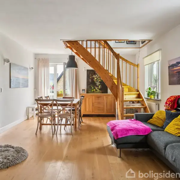 Grå sofa med farverige puder står i dagligstue med trætrappe. Rundt spisebord med stole, lysindfald fra vinduer. Væggene har kunstværker, og gulvet er lys træ.