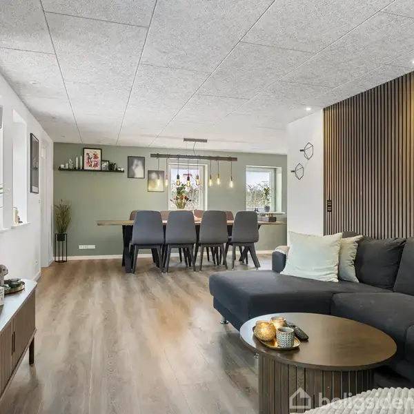 En moderne stue med en stor, mørkegrå sofa ved siden af et rundt sofabord. I baggrunden ses en spiseplads med et langt bord og otte stole.