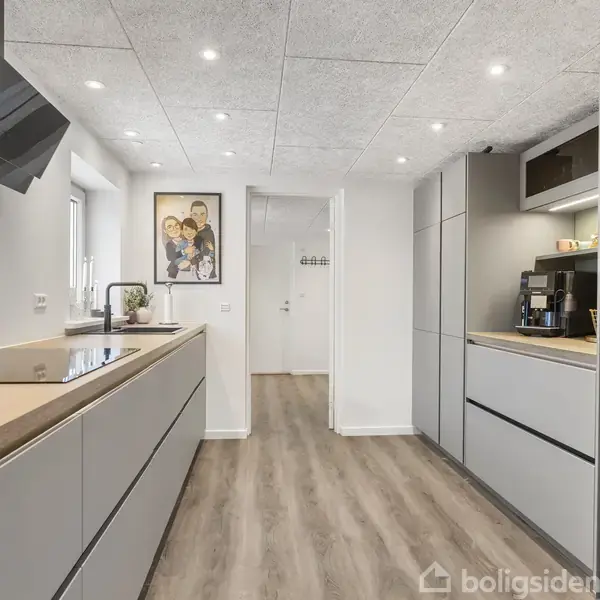 Køkken med grå skabsfronter og træbordplade, kaffemaskine og køleskab; belyst af loftspot. Billede på væggen. Trægulv og hvide vægge skaber en moderne atmosfære.