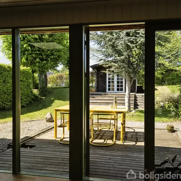 En gul udendørs spisegruppe står på en træterrasse, omgivet af en grøn have med træer og buske. I baggrunden ses et lille sort-hvidt skur.
