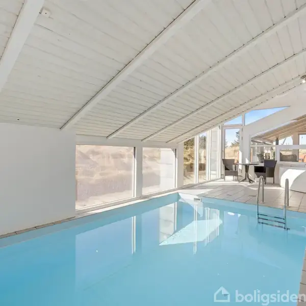 Indendørs swimmingpool med klart blåt vand, placeret i et rum med hvide vægge og loft, store vinduer giver udsigt til en udendørs gårdhave. En trappe fører ned i vandet.
