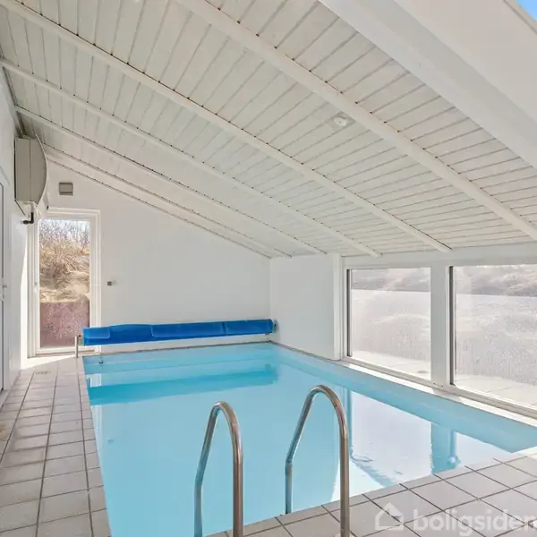 En indendørs swimmingpool med metalstige står i et lyst rum med skråt hvidt paneltag og store vinduer. Fliser dækker gulvet, og en dør fører til udendørsområdet.
