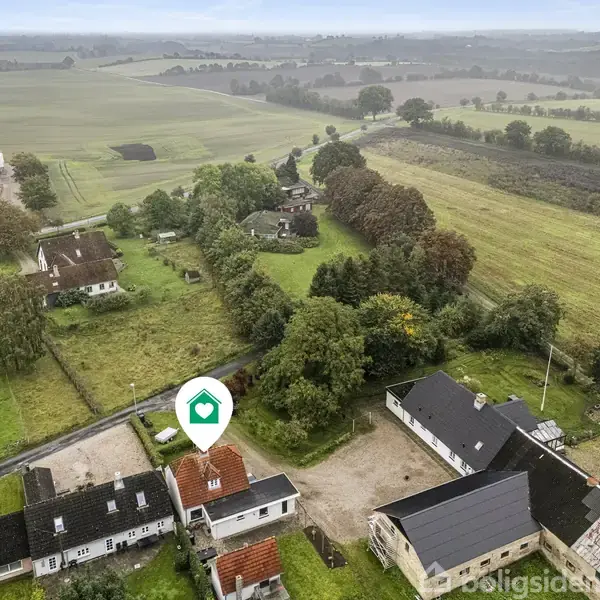 Et hus med en rød tag markeret med et ikon ses i en landlig landsby. Omgivet af grønne marker og andre huse under en overskyet himmel.