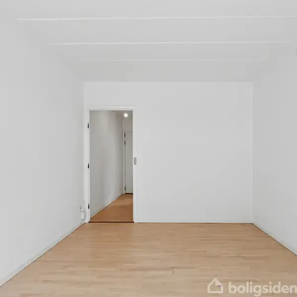 Et tomt rum med hvide vægge og lyst trægulv; en åben dør fører til en gang med yderligere naturligt lys.