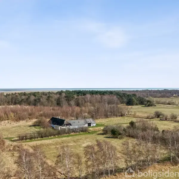 En stor hvid bygning står stille omgivet af åbne marker og spredte træer under en klar blå himmel. Fjern udsigt viser skov og landbrug.