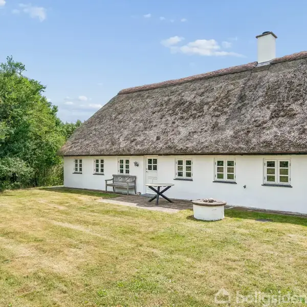 Stråtækt hus med hvid facade står stille ved en terrasse med bord og bænk. Omgivet af en velplejet græsplæne og træer i baggrunden.