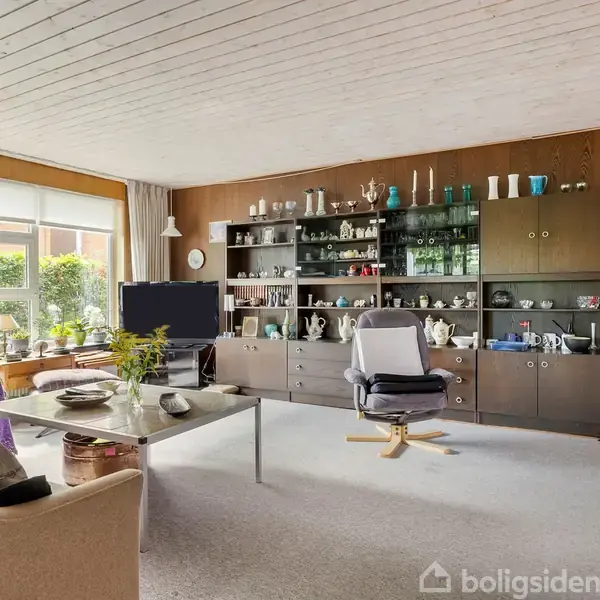 Stue med vitrineskab fyldt med porcelæn og glas, tv på lavt bord, komfortabel stol foran, store vinduer med udsigt til have, og tæppe på gulvet.