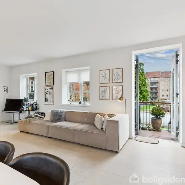 Grå sofa står mod en væg med kunst, der fører til en åben altandør. Rummet har lyse vægge, et køkkenhjørne, spiseplads og udsigt til en bygning udenfor.