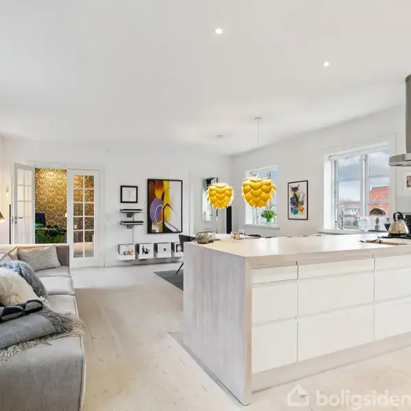 Et moderne køkken-alrum har en stor køkkenø med frugtskål, er omgivet af sofaer og kunstværker på væggene. Store vinduer giver udsigt til bygninger udenfor.