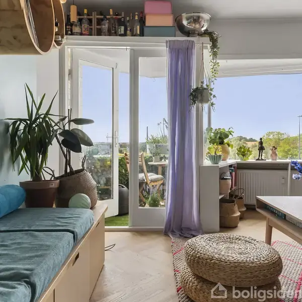 Stue med blå sofa, plante på bord og flettet taburet, åben dør til balkon med planter. Lys træparket, bøger og dekorative genstande øverst. Vindue med udsigt til himmel.