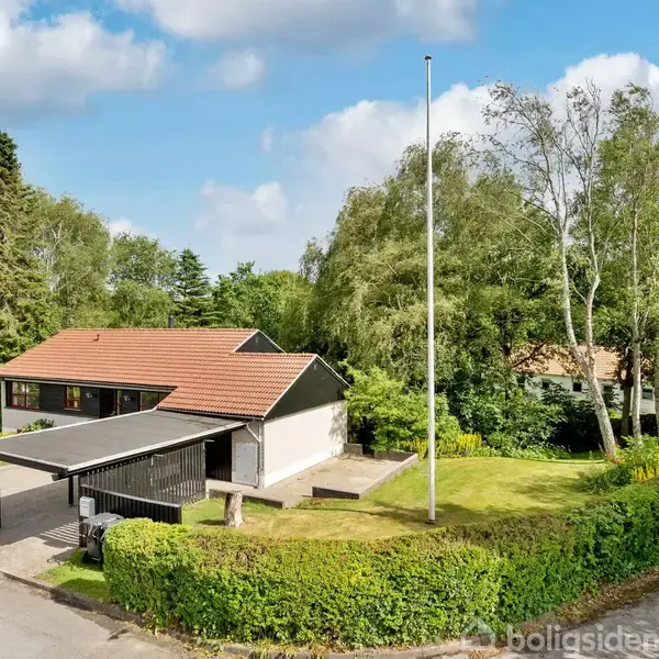 Huset er en hvidbygget villa med rødt tag, placeret ved en vej, med en carport og flagstang. Det er omgivet af en grøn have med buske og træer.