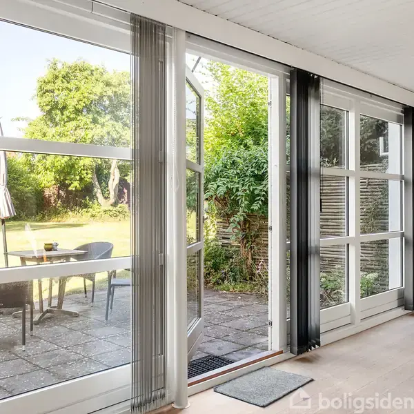 Glasdør åbner ud til terrasse med bord og stole; udenfor er en frodig have med træer. Gulv inde i rummet er lyst, og der er en radiator til højre.