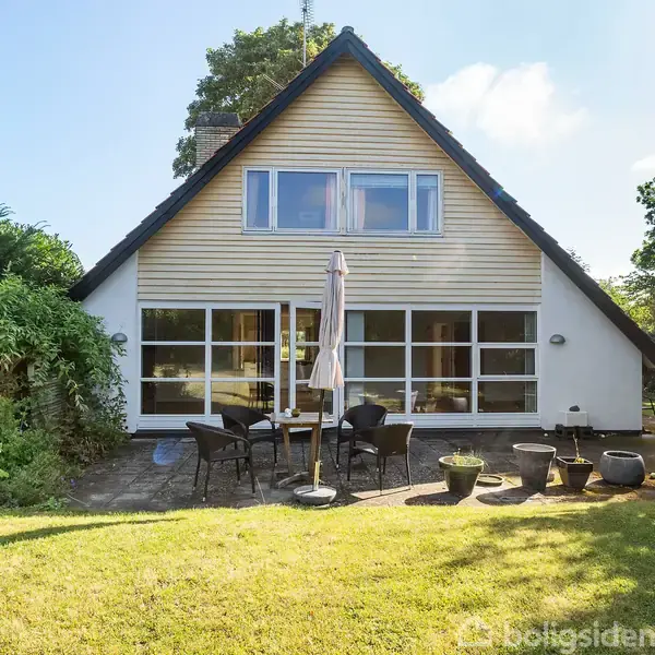 Et træhus med stort vinduesparti og spidst tag står i en have. Foran huset er et terrasseområde med havemøbler og blomsterkrukker. Omgivet af grønne træer og buske.