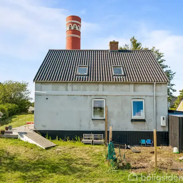 Hus med hvid facade og sort tag står i en have med græsplæne. Til venstre ses en lyskegle og en rød skorsten. Klar blå himmel i baggrunden.