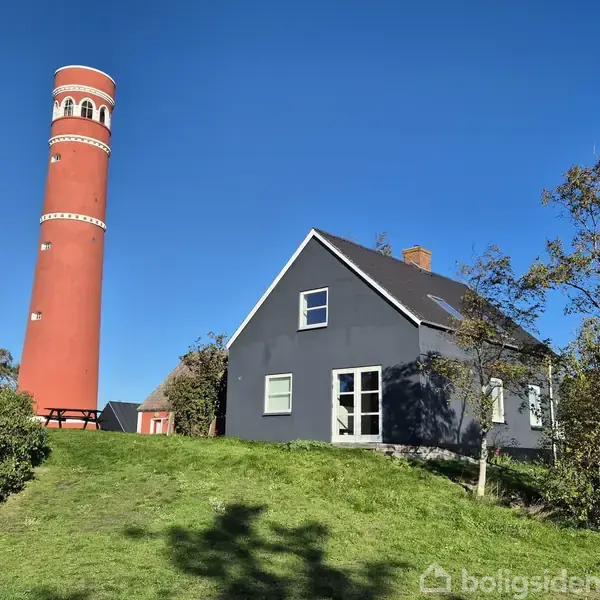 Et rødt fyrtårn står ved siden af et gråt hus med en stejl taghældning. Omgivet af grønne træer og græs under en klar blå himmel.