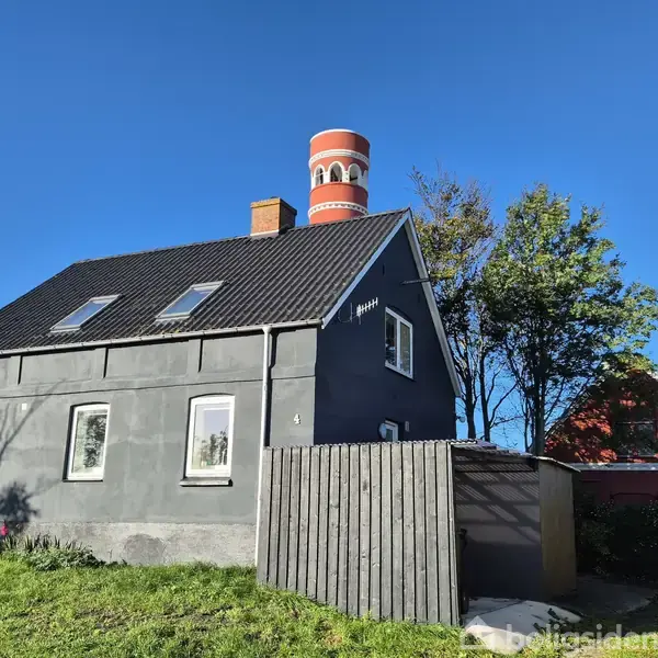 Et gråt hus med to etager står på en grøn græsplæne. Bagved ses et rødt, hvidstribet vandtårn og nogle træer under en klar, blå himmel.