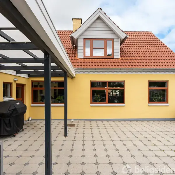 Et gult hus med rødt tag og kvist, set fra en flisebelagt terrasse med havemøbler og en grill. Terrasseoverdækning af glas og metal.