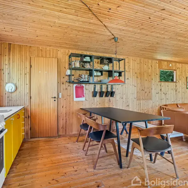 Køkken-alrum med træpaneler på vægge og loft. Sort spisebord med stole, køkkenområde med gule skabe og hvidevarer, rød lampe. Sofa i baggrunden. Vinduer viser grønne omgivelser udenfor.