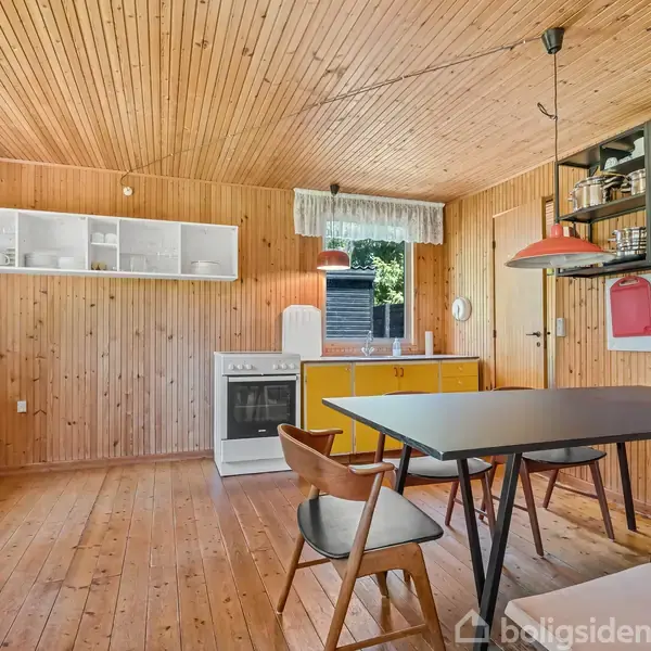 Trækøkken med spisebord og stole. Vægge og lofter har lyse træpaneler. Hvidevarer inkluderer komfur og køleskab. Vindue med gardin lader naturligt lys ind. Hylder med glas og service. Ingen tekst fundet.