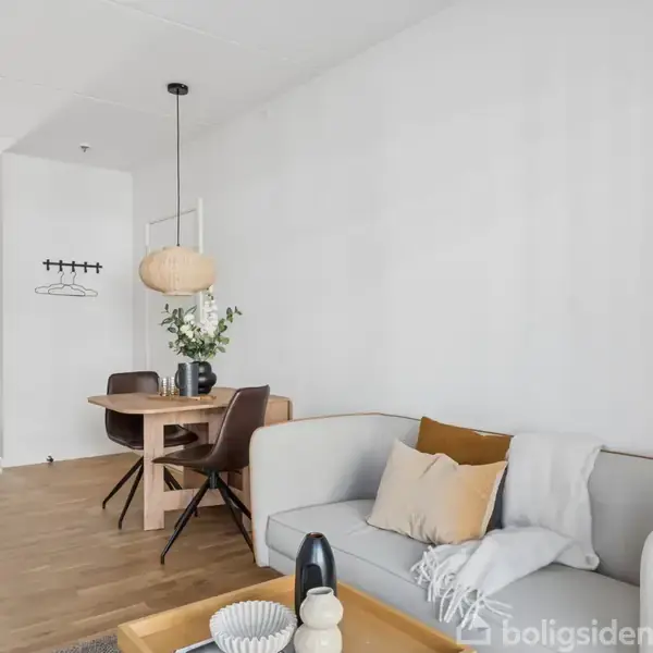 Sofa med puder placeret i en stue ved siden af et rundt spisebord med stole og en hængende lampe. Rummet har hvidmalede vægge og trægulve. En plante står i baggrunden.