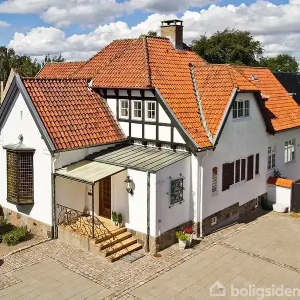 Et hvidt hus med rødt tegltag står i en solrig have omgivet af grønne træer. Huset har en udendørs trappe ved hoveddøren og en mønstret stenbelægning foran.