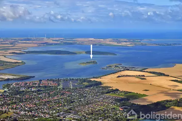 Ø er markeret med en pil, beliggende i en stor sø med omkringliggende landbrugsjord og en by. Der er spredte skyer på himlen. Markante landskabsområder ses omkring.