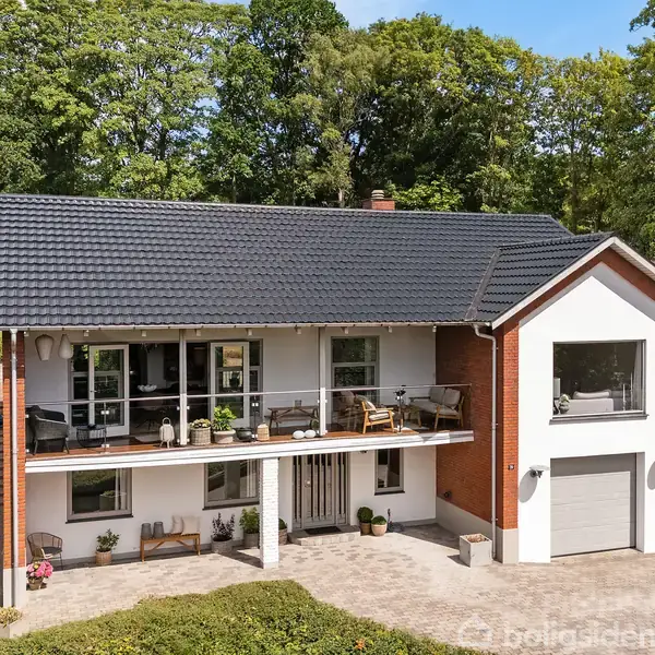 Hus med mørkt tegltag og balkon, omgivet af frodig grøn beplantning, har en garage i underetagen. Pålidelig og rummelig, beliggende i et naturskønt område.