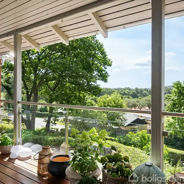 Balkon med trædæk er fyldt med planter i krukker. Den er udstyret med glaspaneler og vender ud mod et frodigt, grønt landskab med træer og blå himmel i baggrunden.