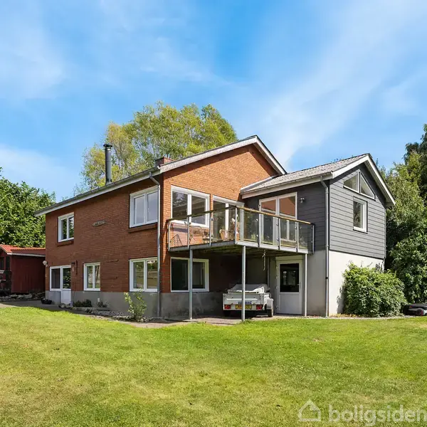To-etagers hus med balkon, omkranset af en grøn have med træer. Huset har brune murstensvægge og et gråt træpanel, og det står under en blå himmel.