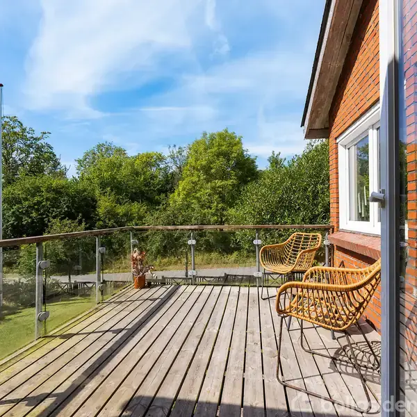 Terrasse med træplankegulv, to stole og glasrækværk. Herfra ses en have med græsplæne og træer. Boligens murstensvæggemøde og en stor glasdør er synlig i højre side.