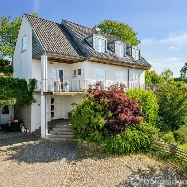 Hvidt hus med skråt sort tag, to etager og altan. Omgivet af frodige træer og buske, stenbelagt indkørsel. Grønt landskab i baggrunden under en klar blå himmel.