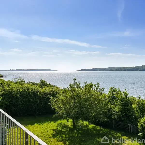 En balkon overlooks en frodig grøn have ved en stille sø med omkringliggende bakker under en klar blå himmel.