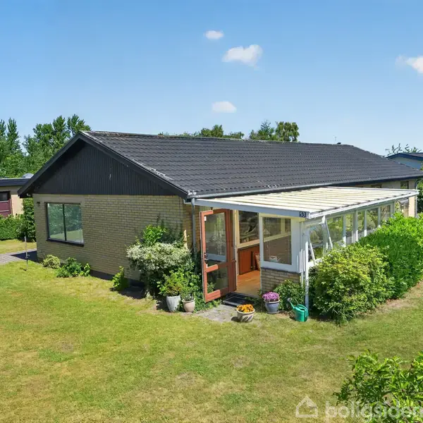 Et enkelt hus med fladt tag er omkranset af en velplejet græsplæne og buske. En overdækket terrasse er synlig. Omgivelserne er et fredeligt boligområde med træer i baggrunden.