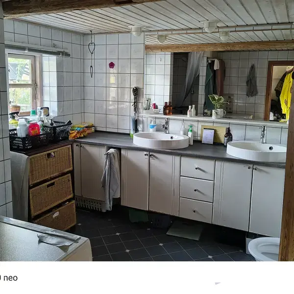 Badeværelse med to håndvaske på en grå bordplade. Skabe nedenunder, spejl og hylder ovenover. Vindue til venstre. Skilt med teksten "Come in, we're open" hænger på døren.