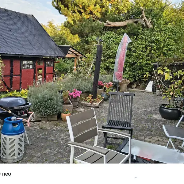 En terrasse med stole og bord står omgivet af grønne planter og blomsterkrukker. En rød bygning med sort tag ses i baggrunden, mens en grill er placeret til venstre.