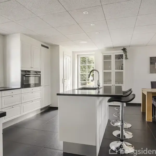 Køkken med hvid indretning og sort bordplade, indeholdende en køkkenø med barstole. Ved siden af står et spisebord med stole i et lyst rum med store vinduer.