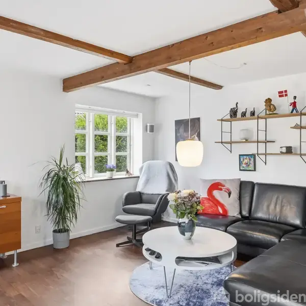Sort L-formet sofa med røde puder står i en lys stue. Rundt sofabord med blomster. Træbjælker i loftet. Vindue, kommode, kunstværk på væggen, og hylder med nipsgenstande.