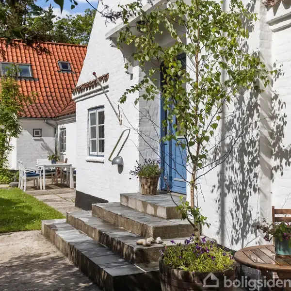 Et hvidt hus med blå dør står i en solfyldt have. En trætrappe fører til døren. Der er en gårdhave med et bord og blomsterkrukke.