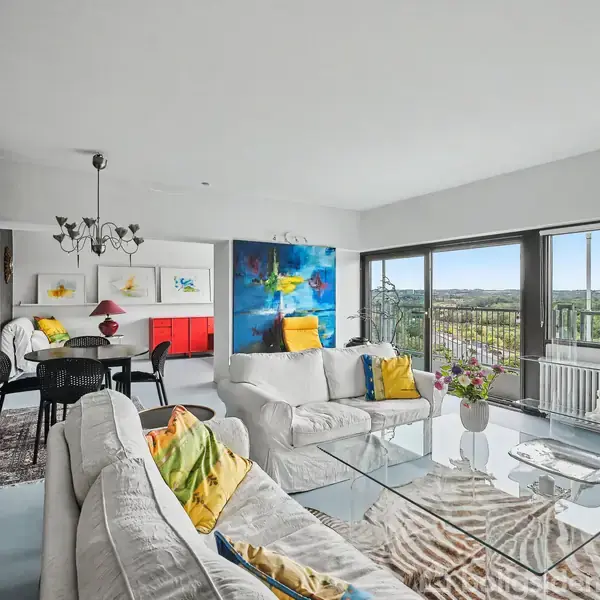Stue med hvide sofaer fyldt med gule puder, placeres foran et glasbord. Store vinduer afslører en grøn udsigt. Til venstre et spisebord med stole under en lysekrone.