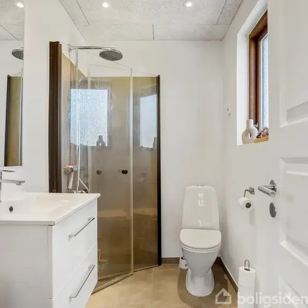 Badeværelse med en brusekabine i glas, hvid håndvask med skuffer, og toilet. Rummet er lyst med et vindue, beige gulv og spotlys i loftet.