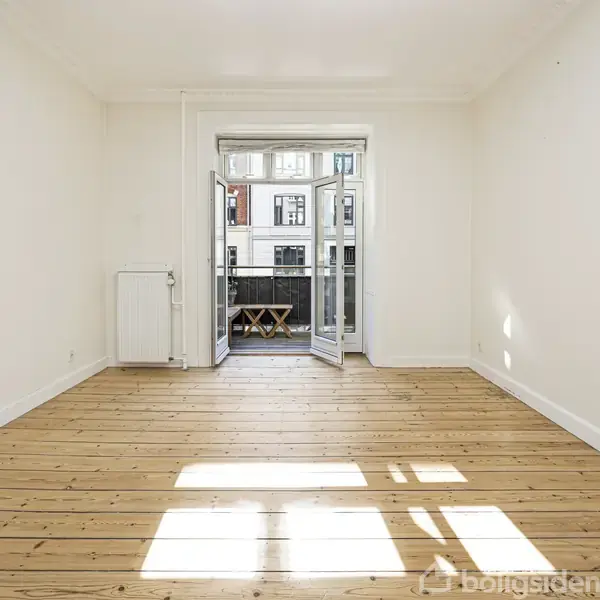 Et lyst rum med trægulv og åbne døre til en balkon. Udenfor ses bygninger. Der er ingen møbler undtagen et lille bord og stole på balkonen.