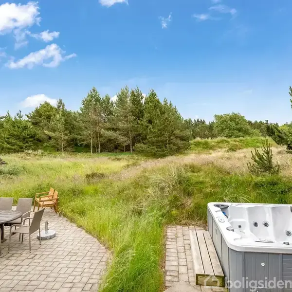 Udendørs spabad står på en flisebelagt terrasse ved siden af et bord med stole. Terrassen grænser op til en frodig natur med græs og træer under en blå himmel.