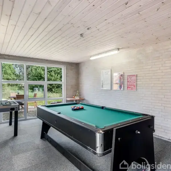 Poolbord står klar til spil i et lyst rum med hvide murstensvægge og store vinduer, som viser en grøn have udenfor. Der er også et bordfodboldspil og poolkøer på væggen.