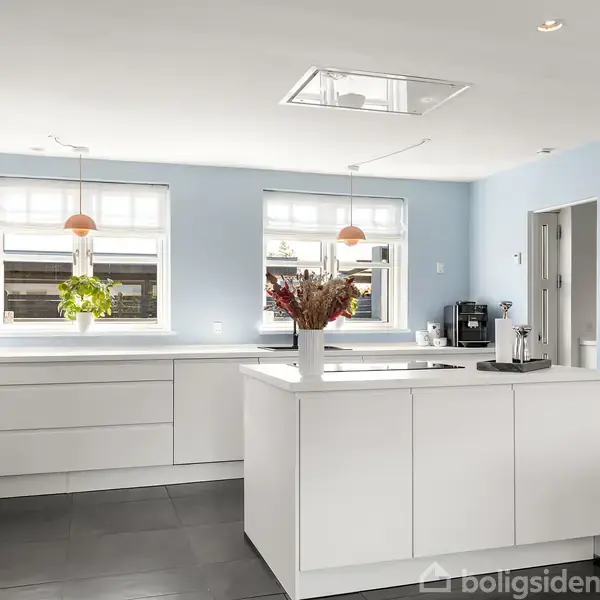 Et hvidt, moderne køkken med en central ø i et lyst rum. To hængende lamper giver lys, og der er planter ved vinduerne. Væggene er lyseblå.