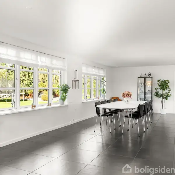 En stort, lyst spisestue med lang, hvid bord og sorte stole midt i rummet. Store vinduer på venstre side slipper naturligt lys ind, og der er indendørs planter og kunst på væggene.