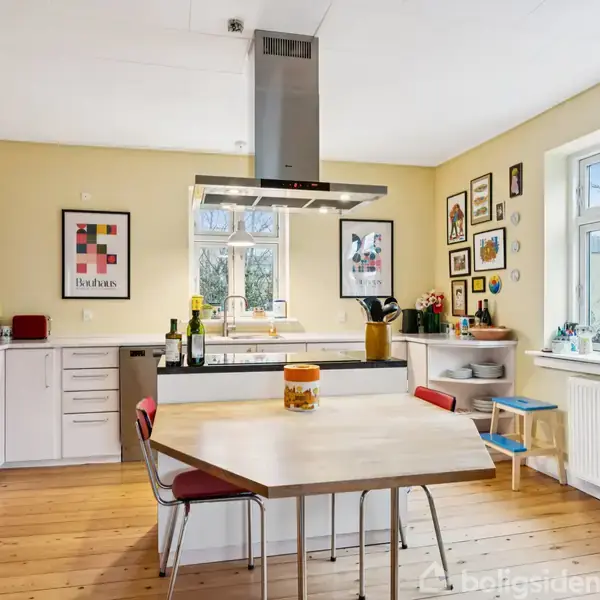 Et køkken med et spisebord i centrum, omgivet af hvide skabe og et stort køleskab til venstre. Vægkunst dekorerer væggene, og store vinduer giver lysindfald.