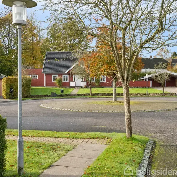 En rød murstensvilla med sort tag står stille i et villakvarter omgivet af træer og en grøn græsplæne. En rundkørsel med et enkelt træ markerer vejens centrum.