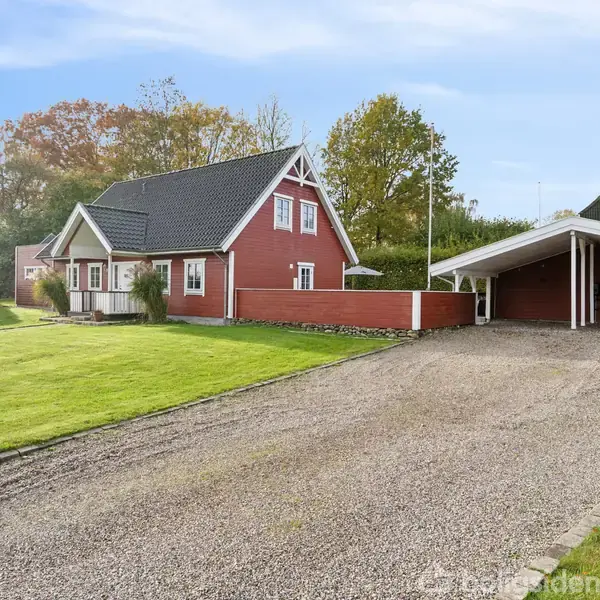 Et rødt hus med hvide detaljer står på en velplejet græsplæne. Ved siden af er der en grusbelagt indkørsel, der fører til en dobbelt carport. Baggrunden viser træer og buske.