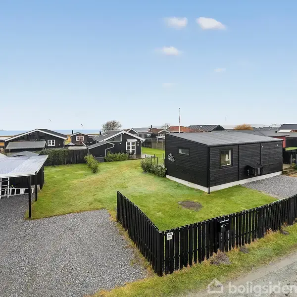 Sort træhus står stille på en grøn græsplæne omgivet af sort hegn. Ved siden af huset er der en grusbelagt indkørsel med en carport. Flere lignende huse ses i baggrunden.